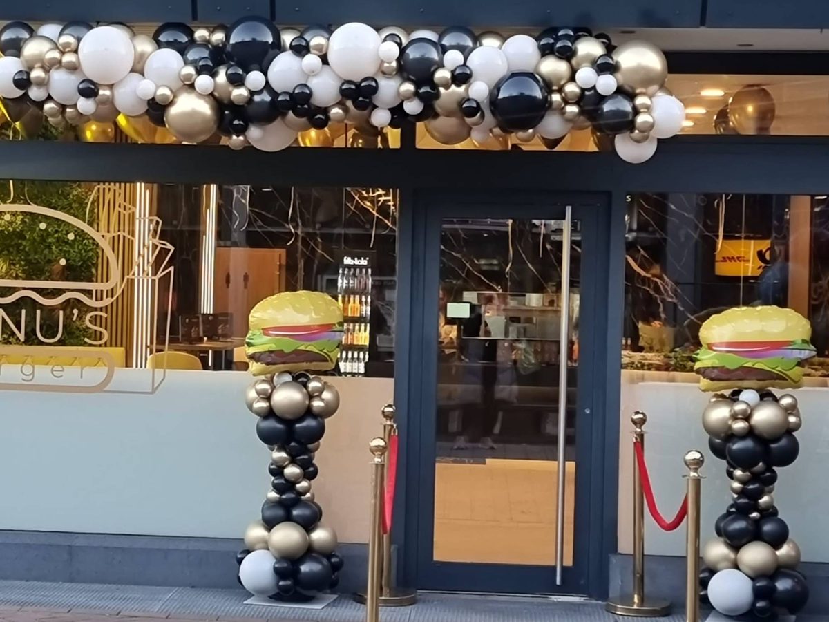 Geschäftseröffnung eines Burger-Restaurants: Organic-Ballongirlande in Schwarz, Gold und Weiß über dem Eingang, zwei Ballonsäulen mit Burger-Folienballons