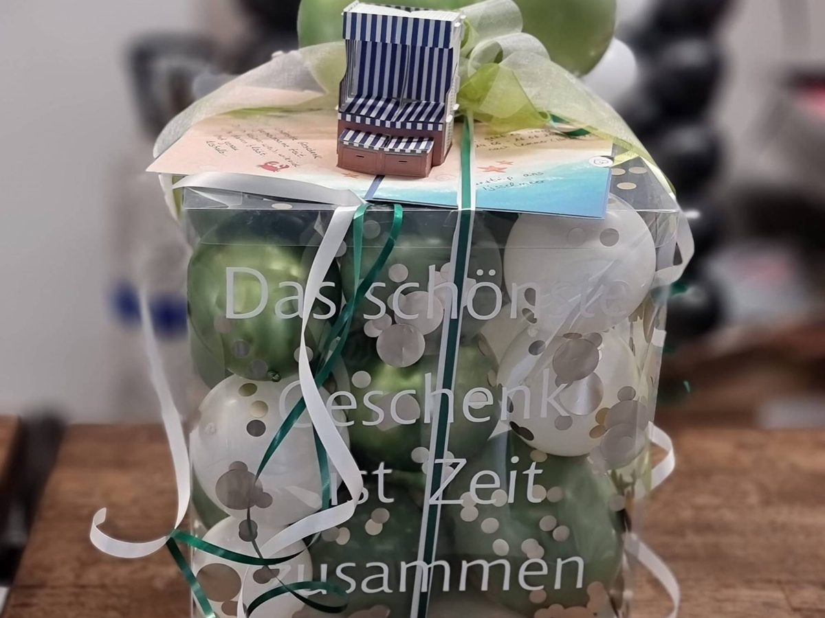 Geschenkbox mit Sichtfenster und Aufdruck „Das schönste Geschenk ist Zeit zusammen“, gefüllt mit Ballons in Grün und Weiß, oben ein Strandkorb aus Papier