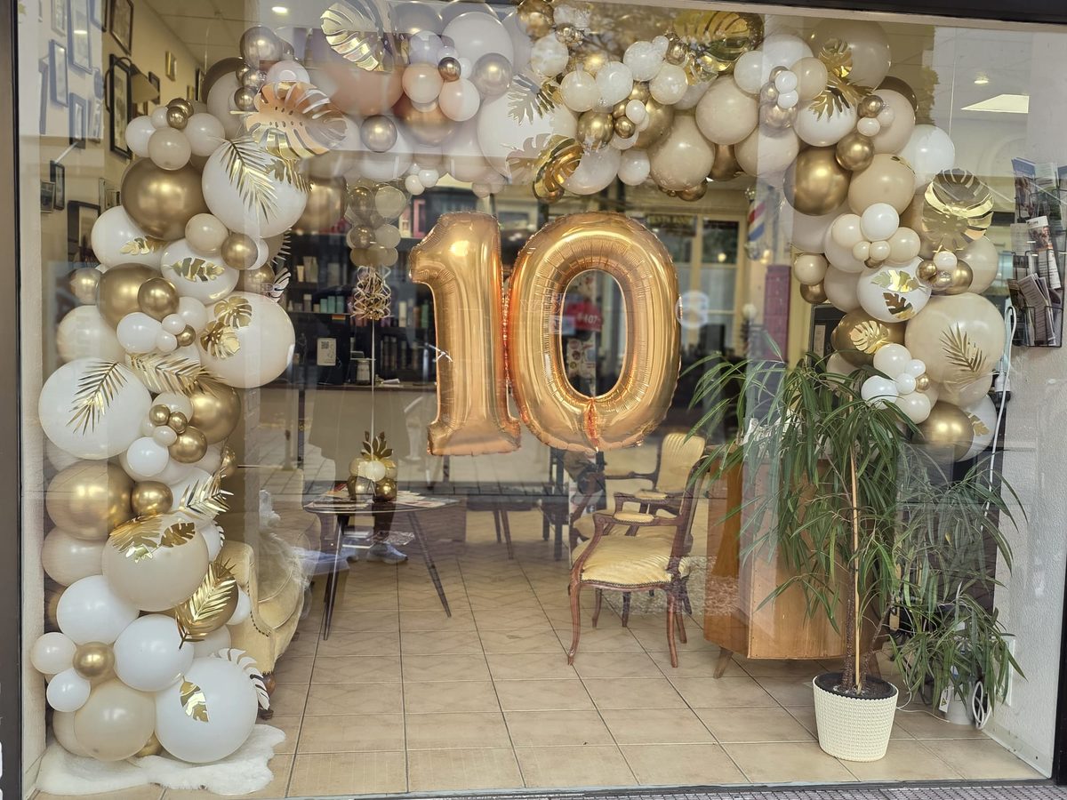 Schaufenster eines Friseursalons mit Ballonbogen in Creme, Beige und Gold, goldenen Palmblättern und großer Folienzahl 10 zum Firmenjubiläum