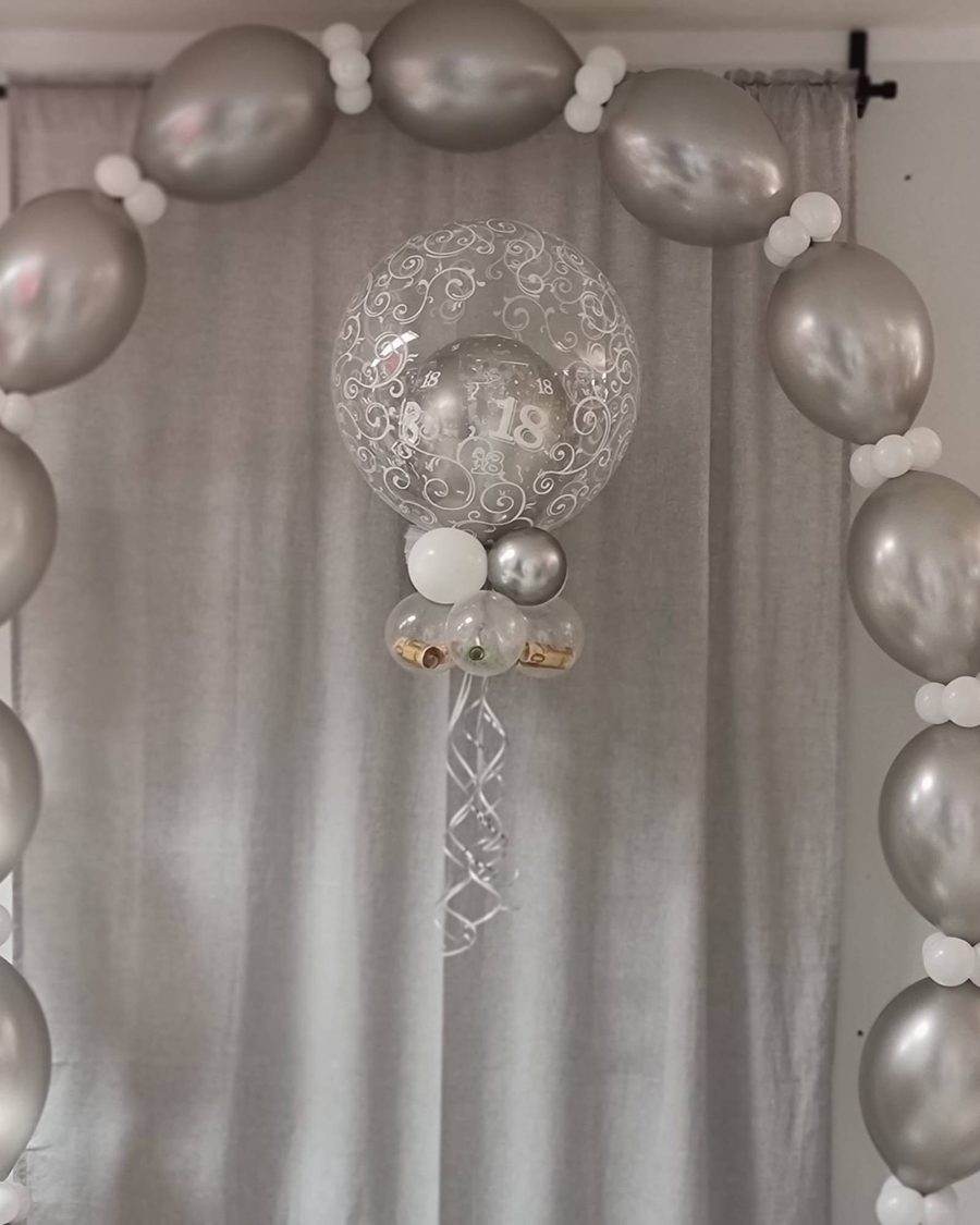 Silberner Ballonbogen, in der Mitte eine Bubble mit Ornament-Print und Konfetti-Zahlen 18, darunter Ballons mit Geldrollen