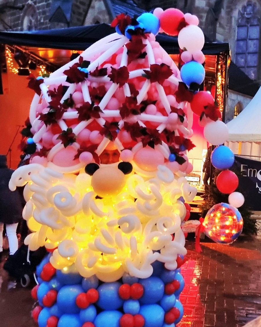 Übergroßer Ballon-Wichtel mit rosa Zipfelmütze, beleuchtetem weißem Bart und blauem Mantel, daneben eine Ballongirlande auf dem Weihnachtsmarkt