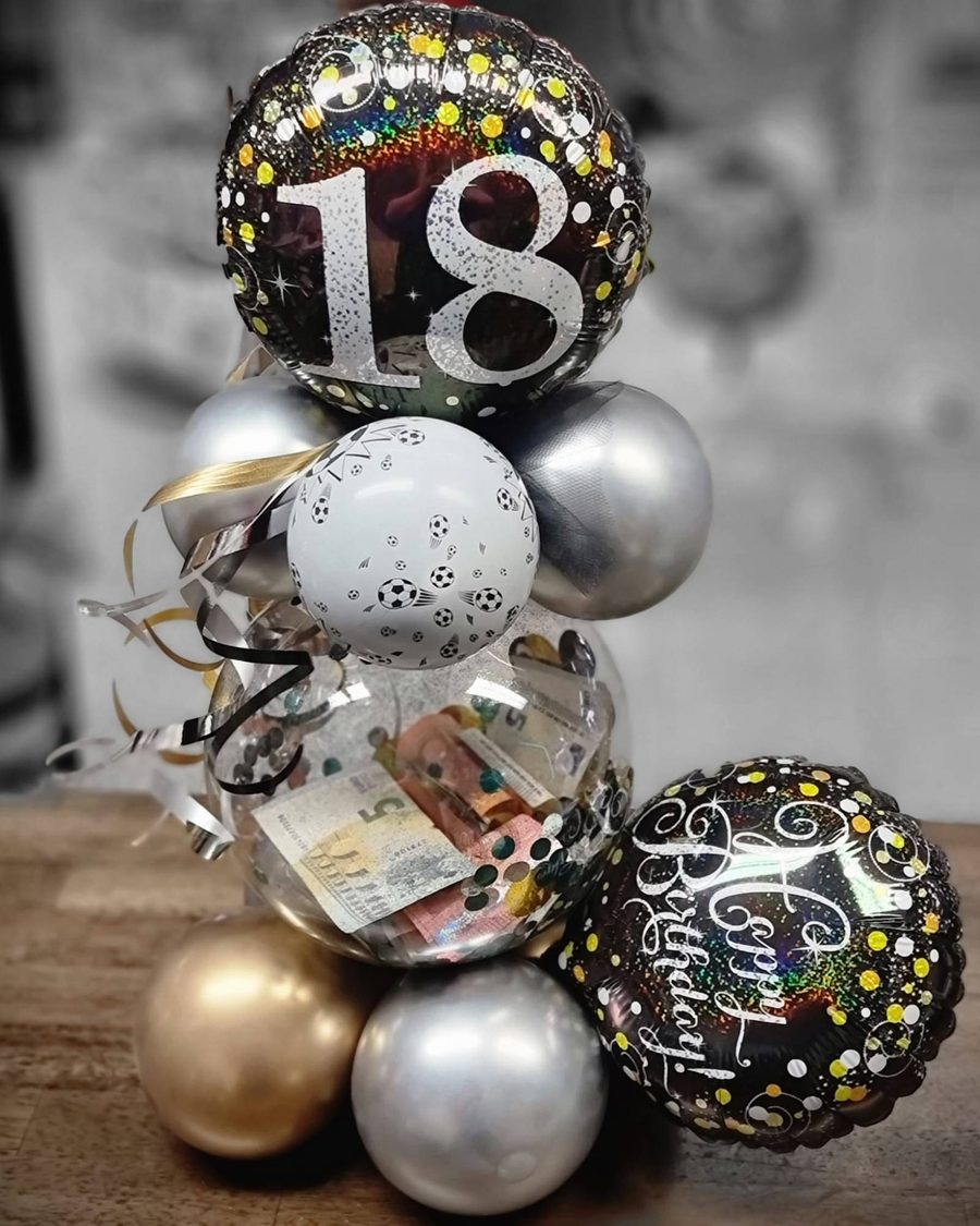 Geldscheine in einem durchsichtigen Ballon, darüber ein 18er-Geburtstagsballon, ein Fußball-Ballon und Ballons in Silber und Gold