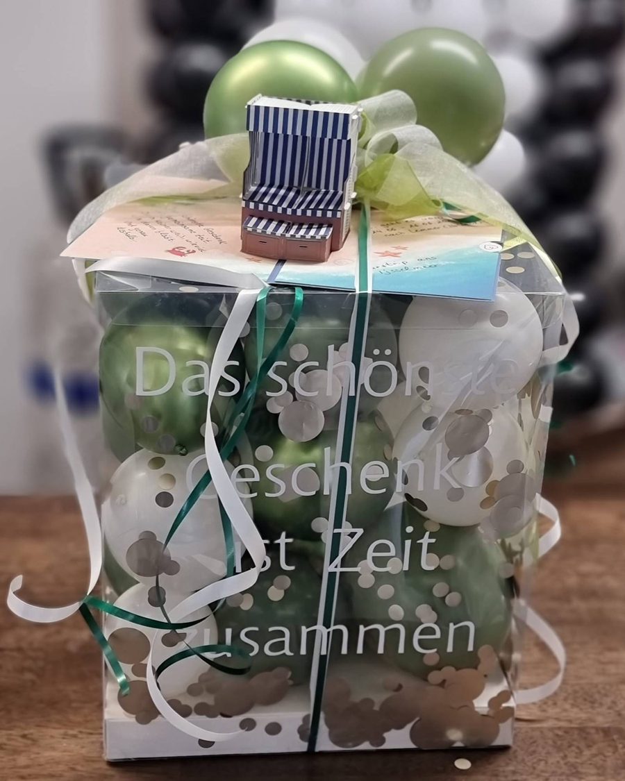Transparente Geschenkbox voller Ballons in Salbeigrün und Weiß mit Schriftzug „Das schönste Geschenk ist Zeit zusammen“, obenauf ein Papier-Strandkorb und eine Karte